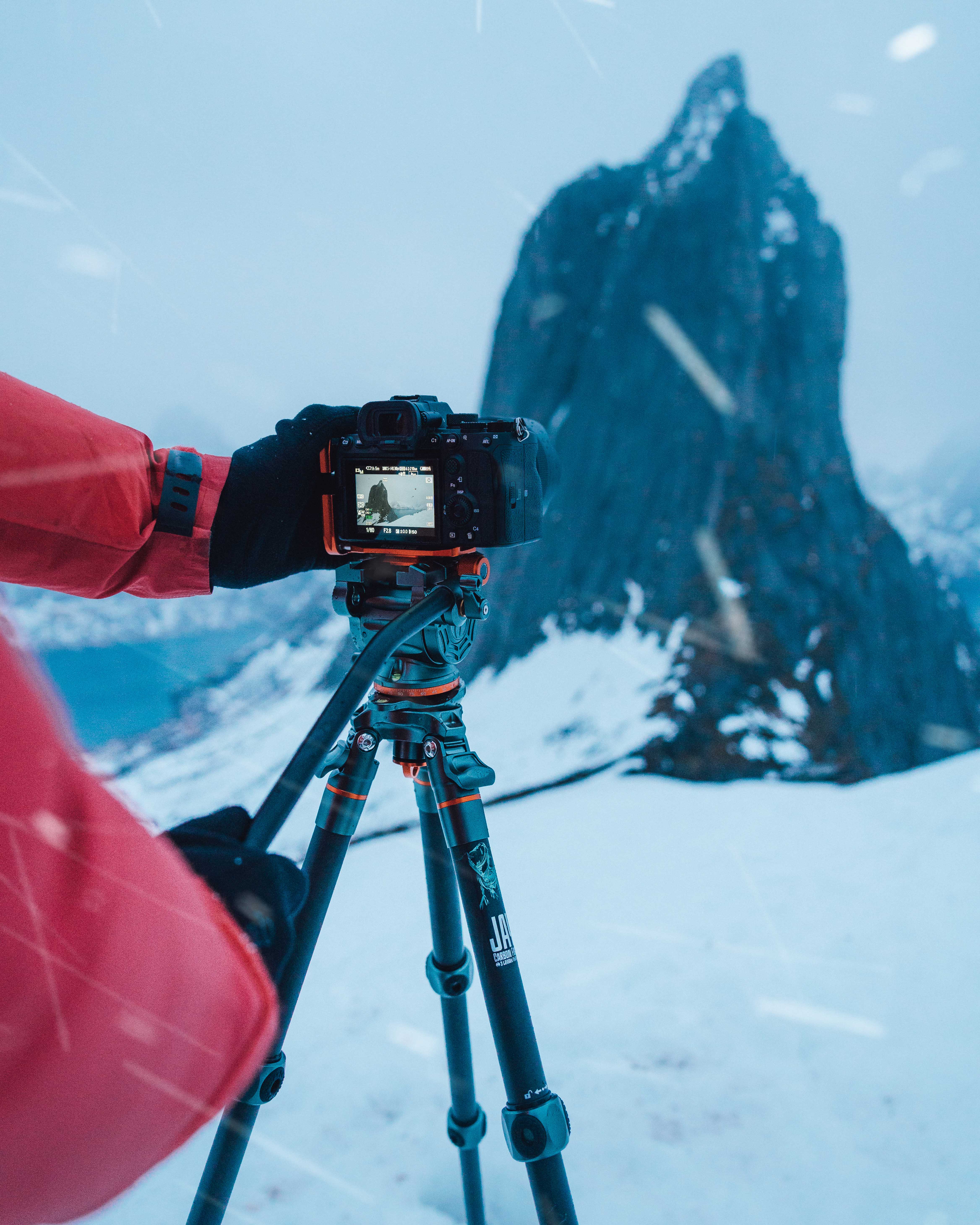 A person in a red jacket takes a tripod photograph of a mountain in the snow